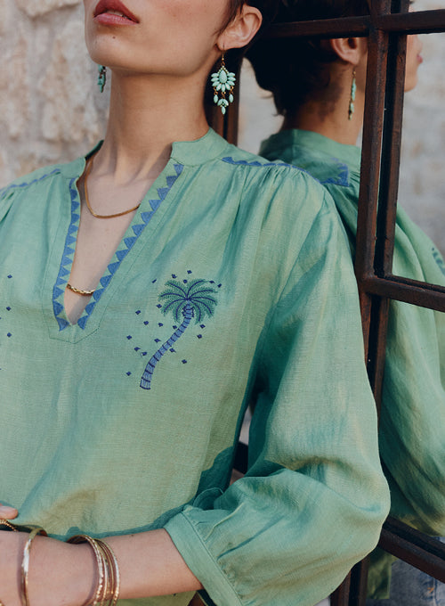 Woman wearing a green embroidered blouse with blue details, standing against a stone wall.