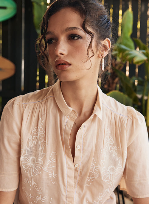 Woman wearing a light pink blouse with floral embroidery against a blurred natural background