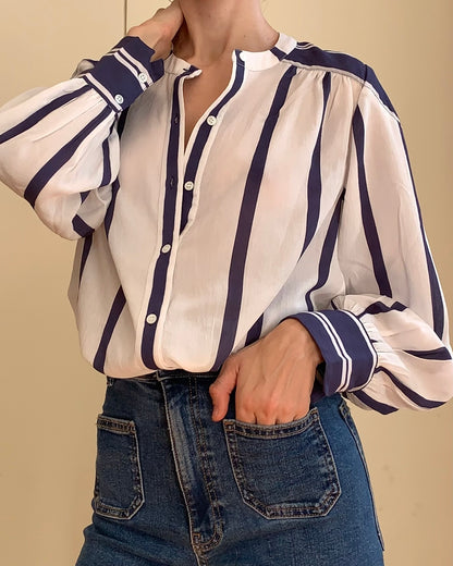 A woman wearing a white long-sleeve blouse with navy blue vertical stripes and button-front placket, paired with blue jeans against a plain beige background.