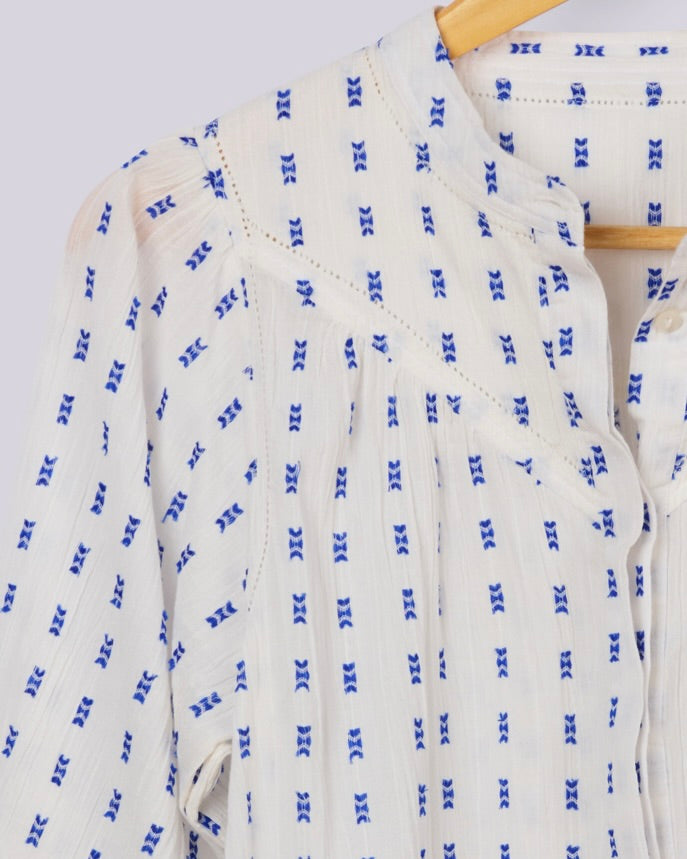 A close-up of a white textured blouse with a blue repeating geometric pattern, hanging on a wooden hanger against a light gray background.