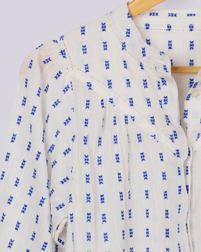 A close-up of a white textured blouse with a blue repeating geometric pattern, hanging on a wooden hanger against a light gray background.