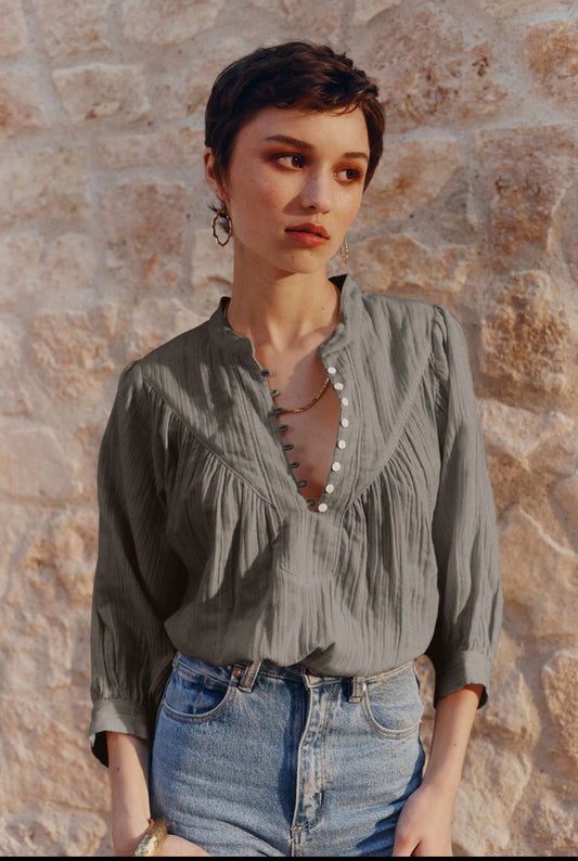 A woman with short dark hair wearing a gray crinkled blouse with a scalloped V-neckline and blue jeans, standing against a rustic stone wall.