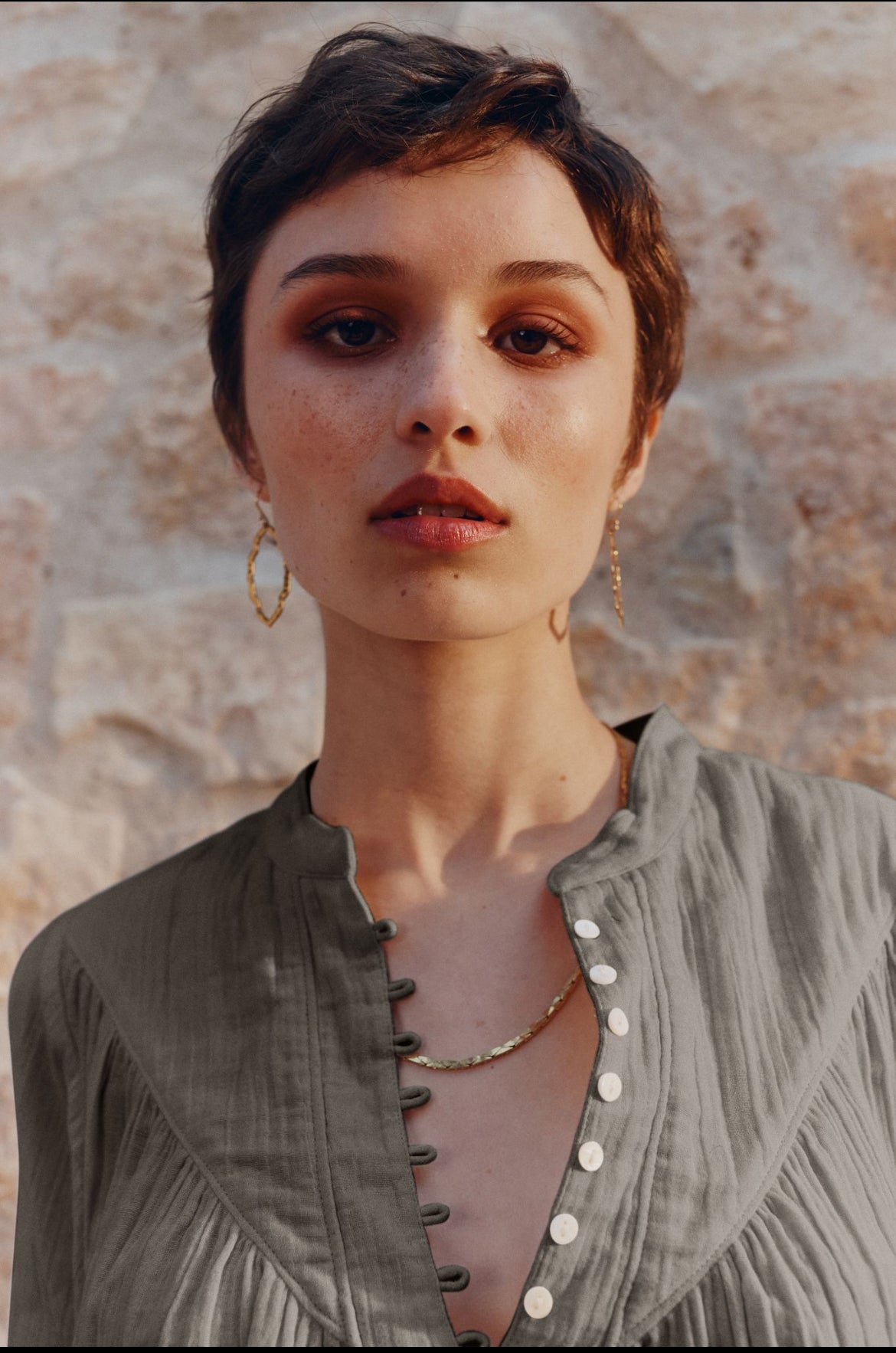 A close-up portrait of a woman with short dark hair wearing a textured sage green top with a buttoned neckline and gold hoop earrings, set against a rustic stone wall background.