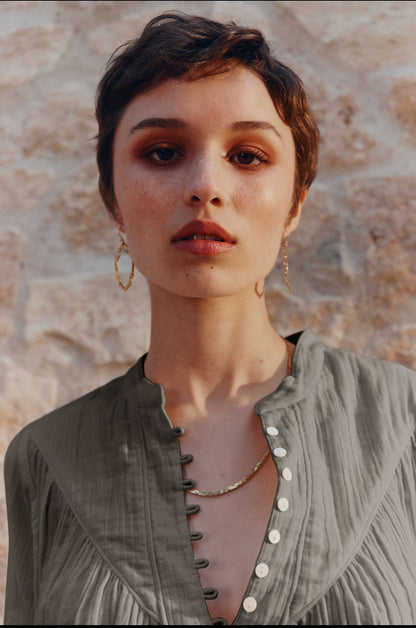 A close-up portrait of a woman with short dark hair wearing a textured sage green top with a buttoned neckline and gold hoop earrings, set against a rustic stone wall background.