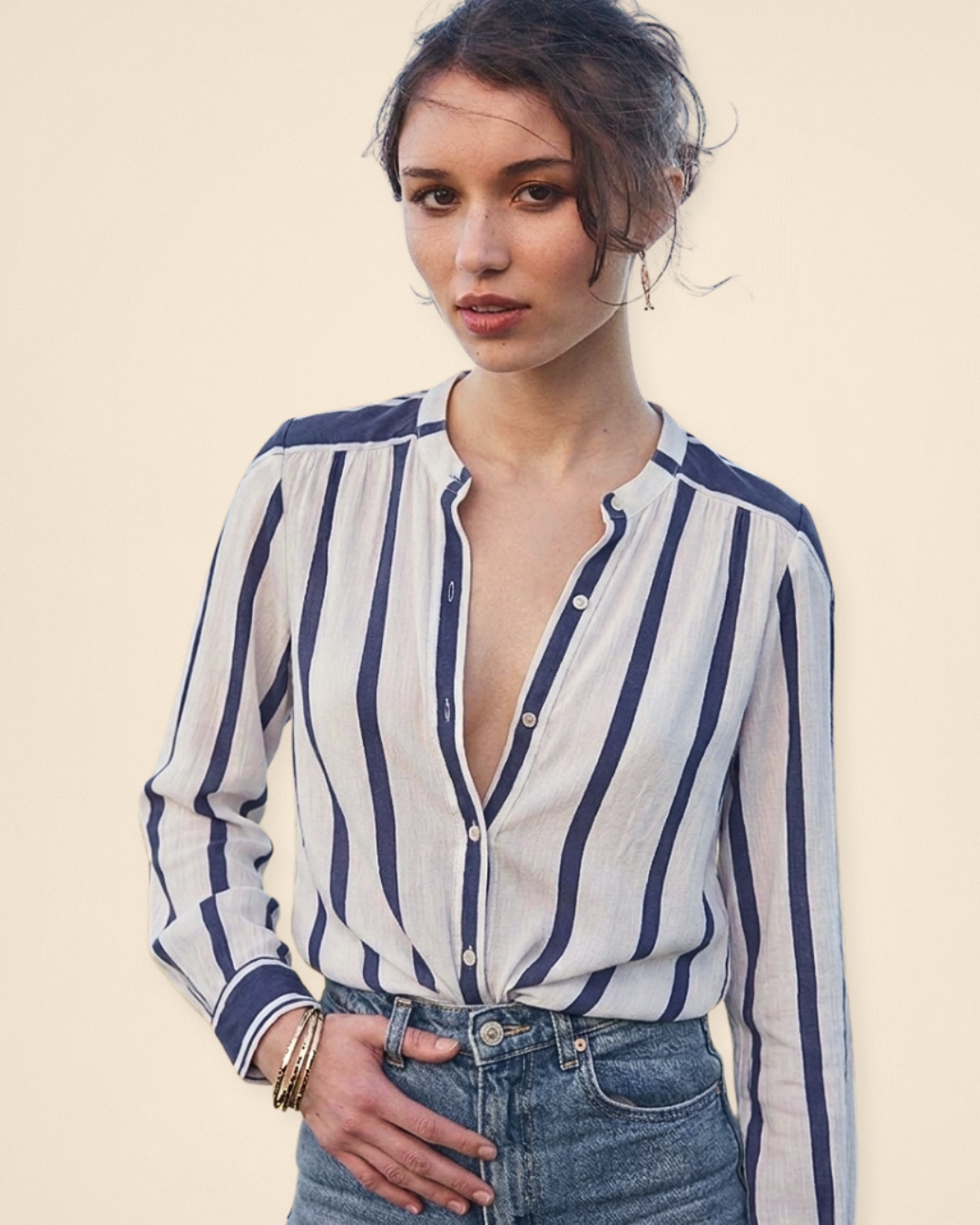 A woman wearing a long-sleeve button-front blouse with vertical navy blue stripes on a white base, paired with blue jeans, against a plain beige studio background.