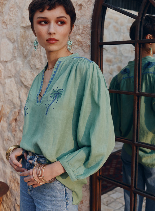 Woman wearing a green embroidered blouse standing against a stone wall.