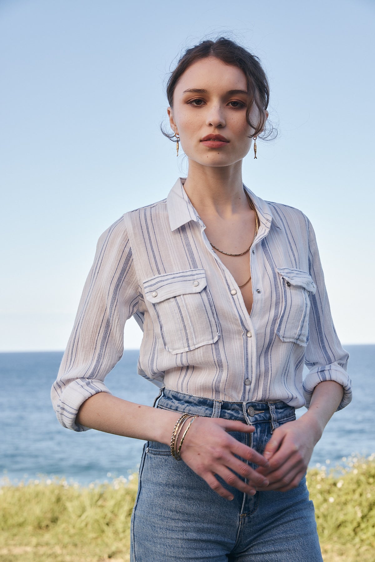 A woman stands outdoors by the ocean wearing a light blue and white vertically striped button-up shirt with two chest pockets, paired with blue jeans.