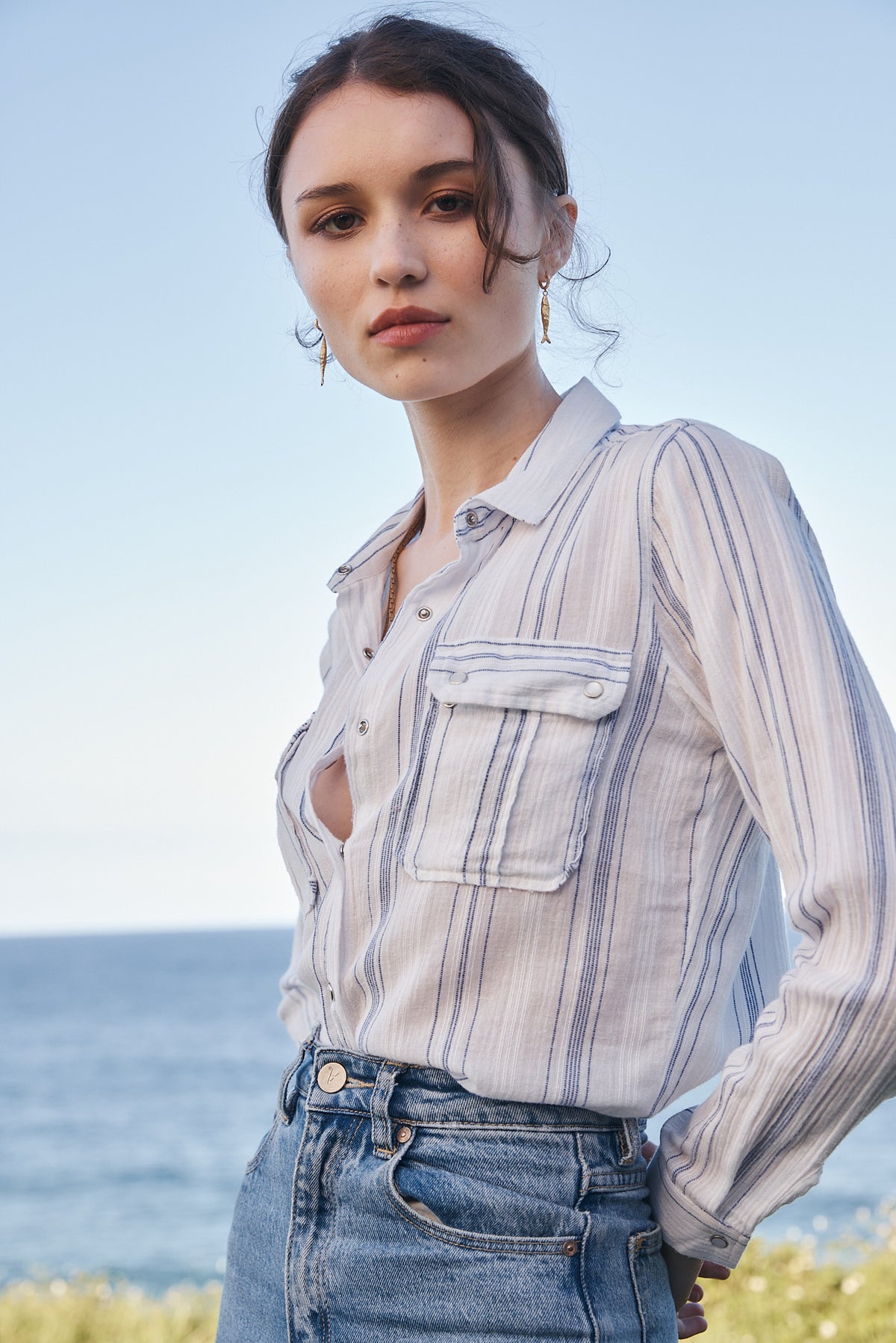 A woman wearing a light beige and blue vertically striped button-up shirt with blue jeans, photographed outdoors against a soft-focus background of the sea and sky.
