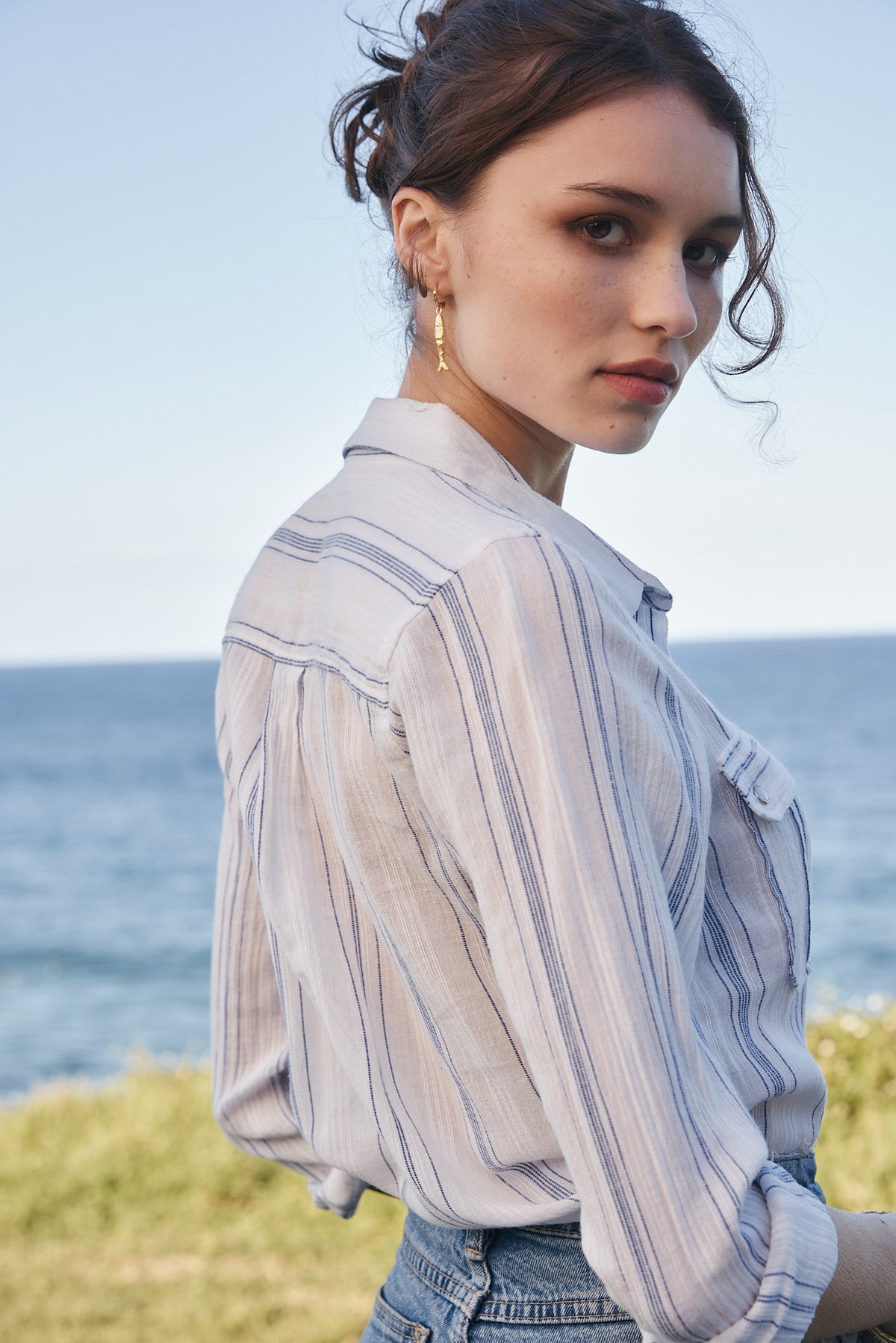 A woman wearing a light blue and white vertically striped button-up shirt, photographed outdoors with the ocean and grass in the background.