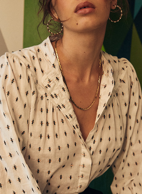 Close-up of a woman wearing a white colored blouse with a small black geometric pattern, layered with a gold chain necklace and gold hoop earrings.
