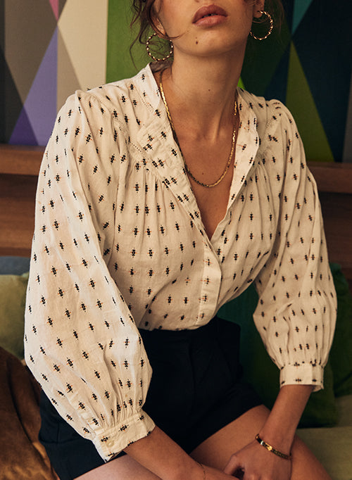 A woman wearing a white long-sleeved blouse with a small black geometric pattern, paired with black shorts. The blouse features a gathered, ruffled collar and voluminous sleeves with elastic cuffs.